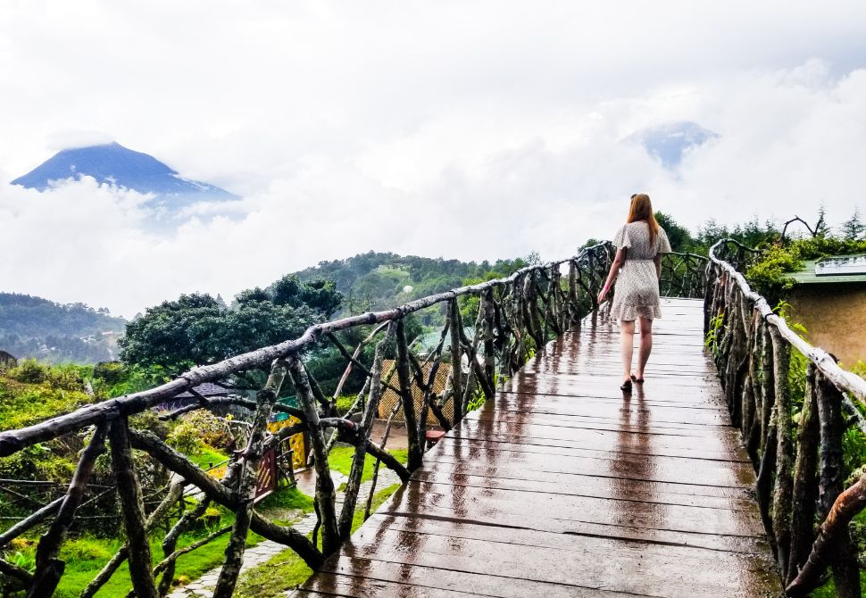 A hobbitenango bridge with wooden railings and a volcano poking out through the clouds in the background
