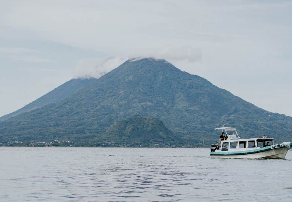 A large volcano on the edge of a large lake with a small white boat floating by - hiking volcanoes in Guatemala